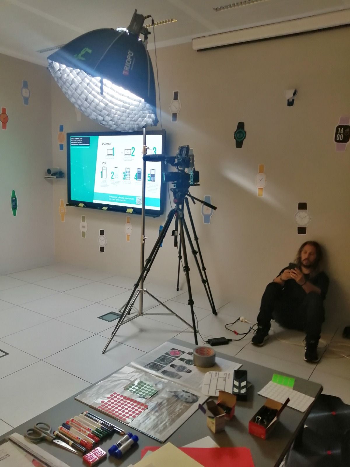 Un set de rodaje: Un hombre se sienta junto a una cámara sobre un trípode, frente a un monitor y una luz de caja de luz. El set está en una habitación con relojes en la pared.