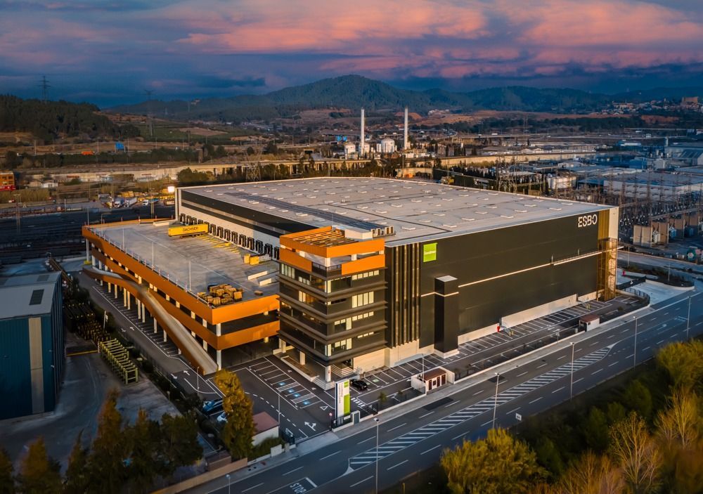 Gran edificio industrial moderno, almacén de ZARA, al atardecer con montañas al fondo. Exterior oscuro con detalles en naranja y negro.