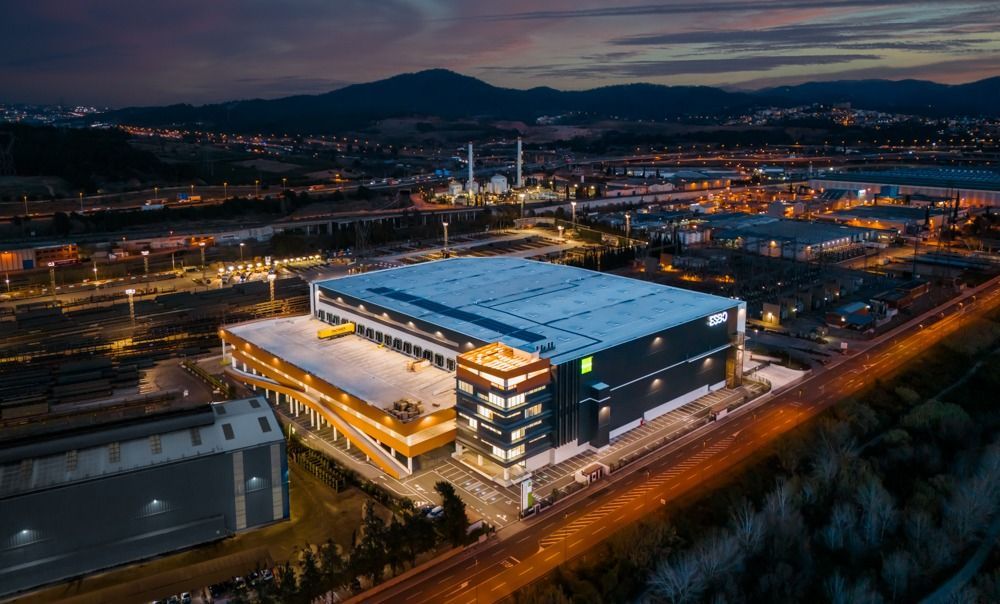 Vista aérea nocturna de un gran edificio industrial con una torre de varios pisos, iluminado con luces brillantes. Al fondo se ven montañas lejanas y las luces de la ciudad.