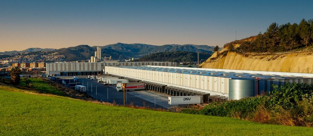 Un gran edificio industrial con un muelle de carga, con un telón de fondo de montañas y un campo de hierba.