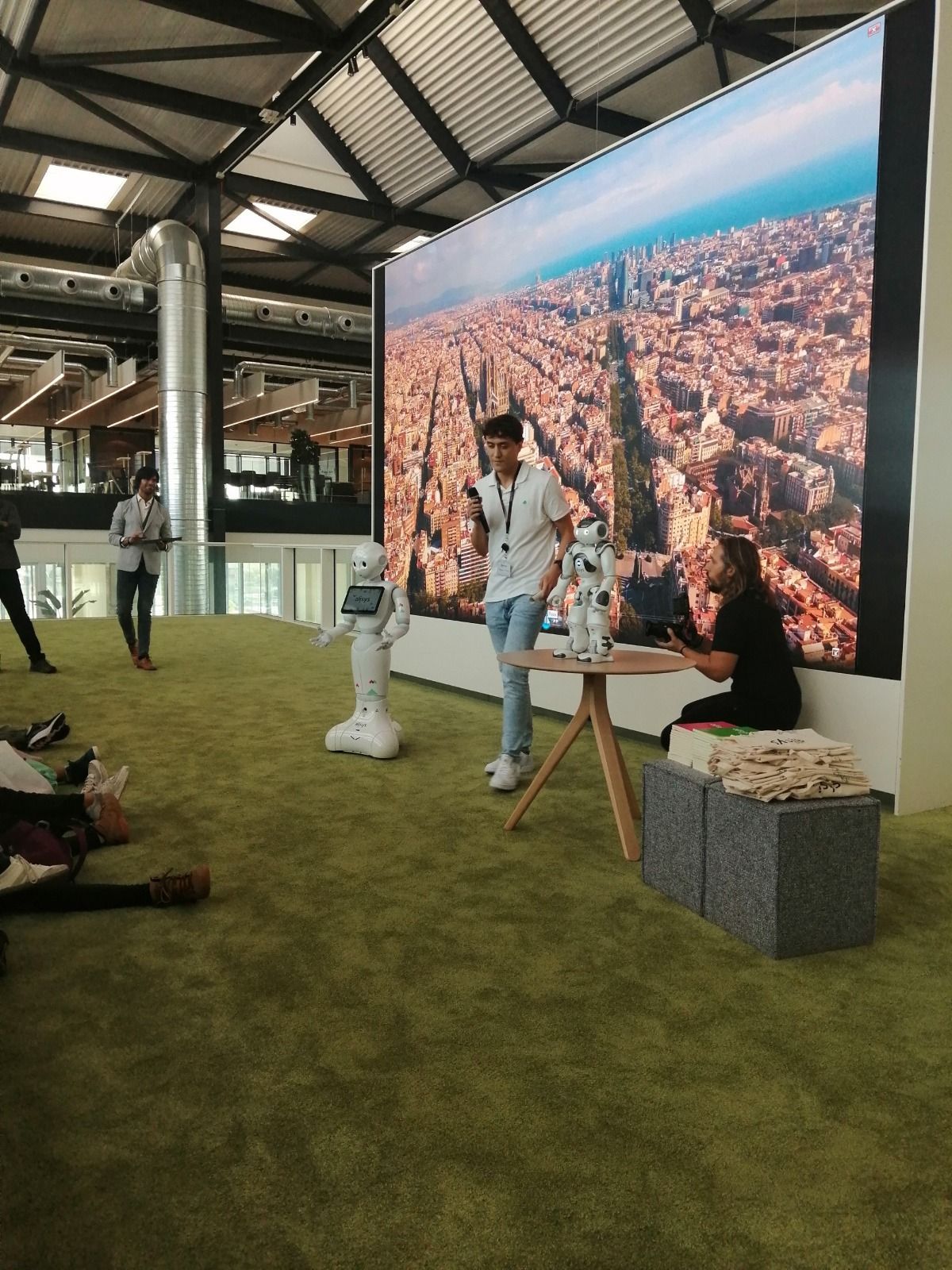 Un hombre con dos robots en un escenario con un paisaje urbano de fondo. La gente se sienta y se para sobre una alfombra verde.