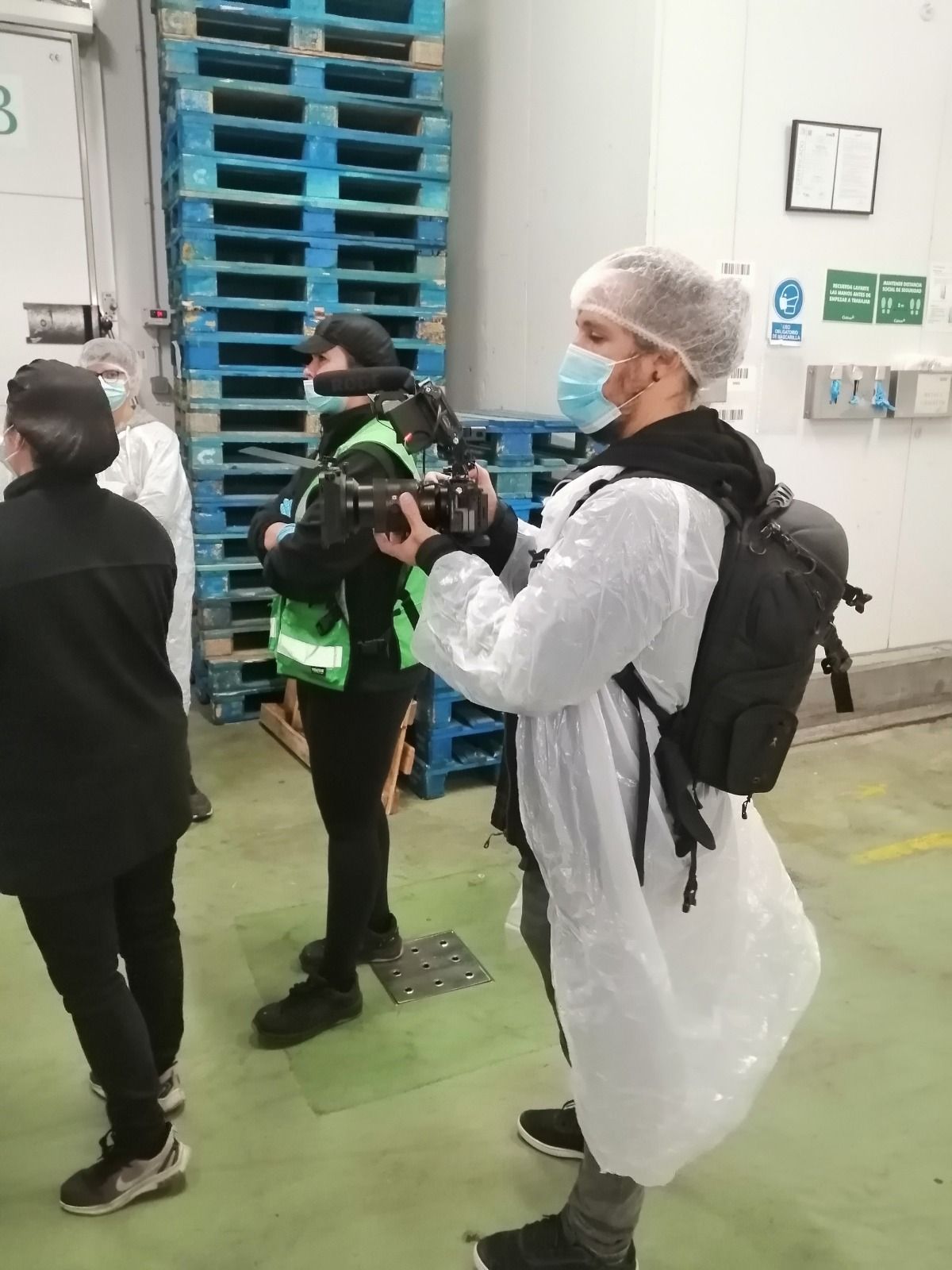 Una persona con traje protector y redecilla graba con una cámara en un almacén. Hay otras personas cerca, incluyendo alguien con chaleco salvavidas.