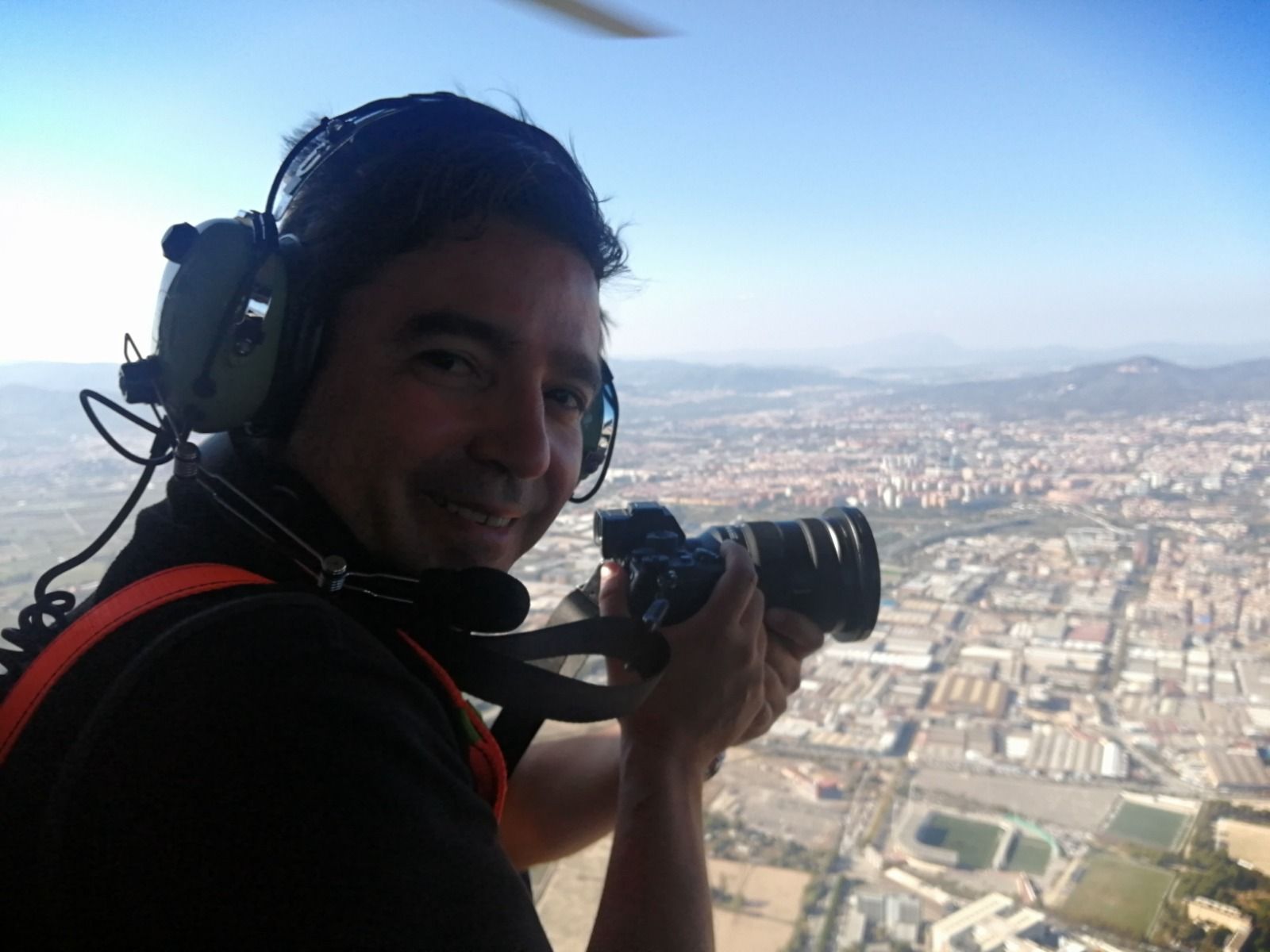 Un hombre con auriculares sonríe mientras sostiene una cámara desde el costado de un helicóptero, capturando una vista aérea de la ciudad.
