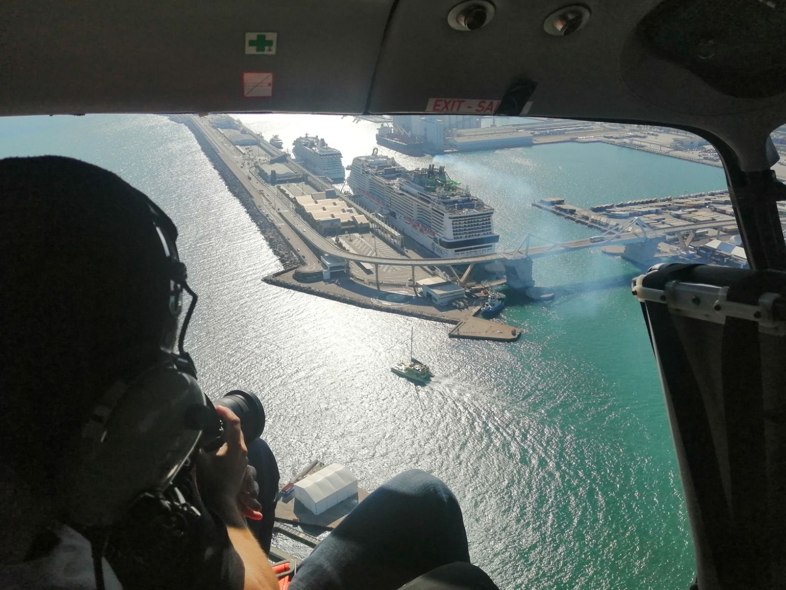 Vista desde la ventana de un helicóptero con vistas a un puerto con un crucero atracado y una persona en primer plano con auriculares.