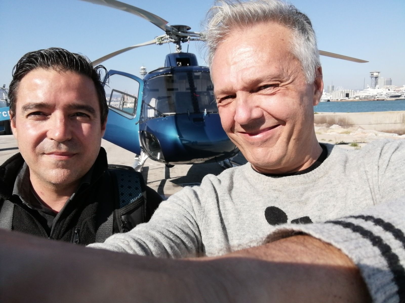 Dos hombres sonriendo, tomándose una selfi frente a un helicóptero azul. El hombre de la derecha tiene el pelo canoso y el de la izquierda, el oscuro. Un entorno soleado al aire libre.