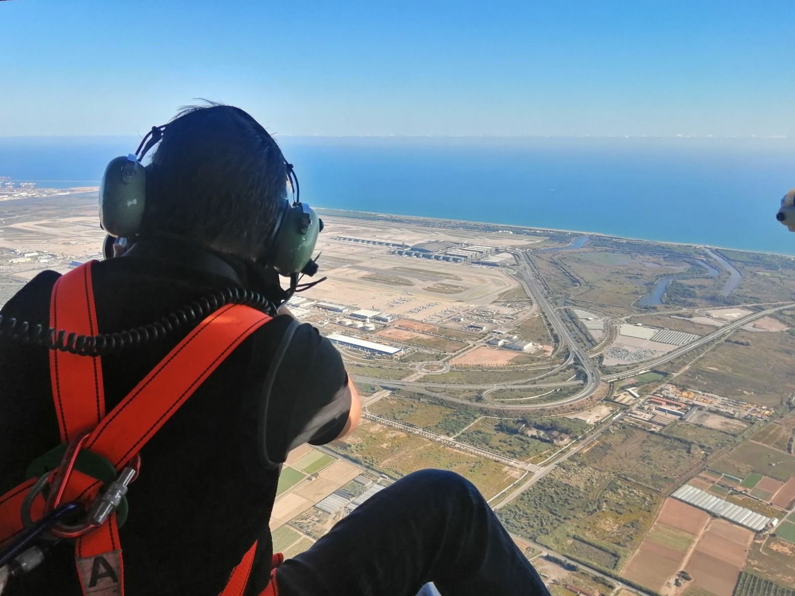 Persona en helicóptero, mirando el paisaje costero con autopista, campos y océano bajo un cielo azul.