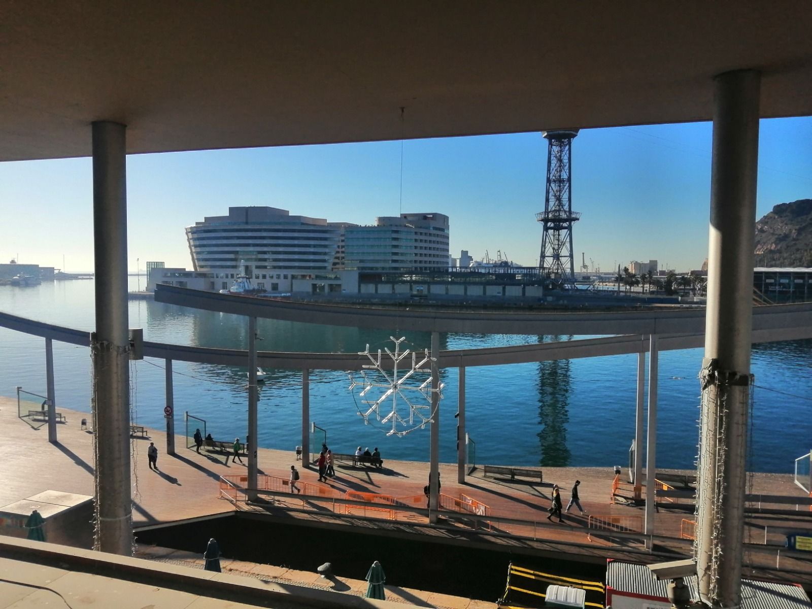 Vista del puerto de Barcelona, España, enmarcada por una estructura moderna con pilares. Al fondo, un gran edificio blanco y el agua. La gente camina por un muelle.