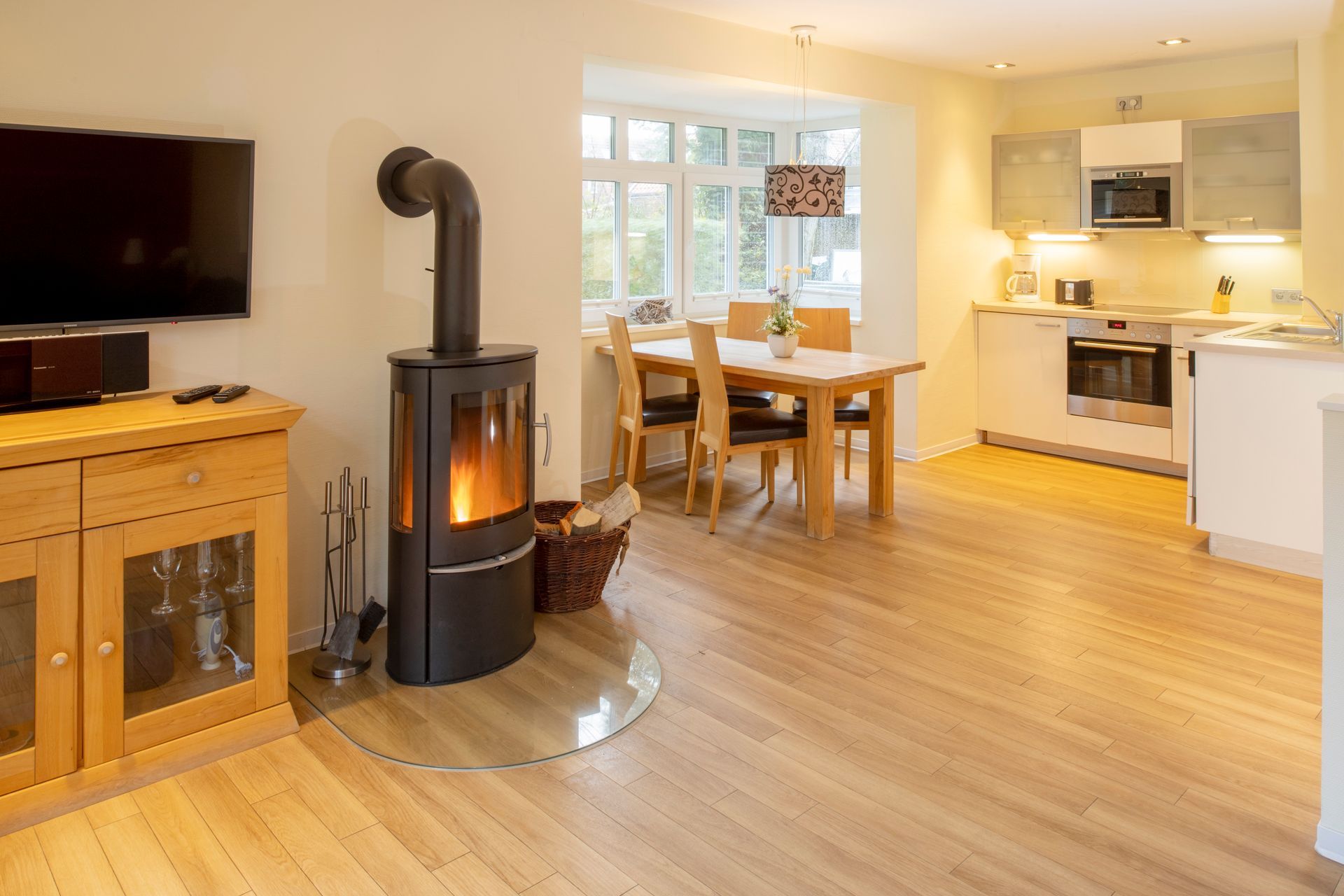 Wohnzimmer mit Kamin, Holzboden, Esstisch und Küche im Hintergrund.