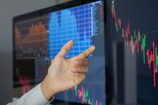 Mano señalando un gráfico de acciones en la pantalla de una computadora. Se muestran barras rojas y verdes, gráficos y datos.