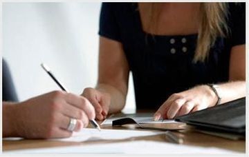 Persona firmando documento en el escritorio, otra persona observando.