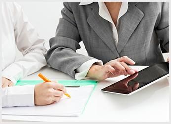 Dos personas en un escritorio: una escribe en papel y la otra usa una tableta. Llevan ropa de oficina y un fondo claro.