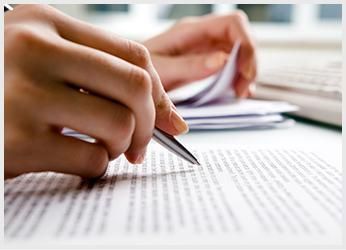 Manos escribiendo con un bolígrafo sobre papel, otros papeles en el fondo.