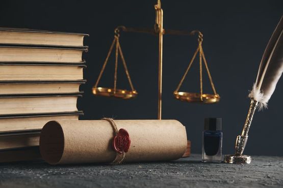 Libros, balanza de la justicia, pergamino con sello, tintero y pluma de ave sobre una superficie oscura.