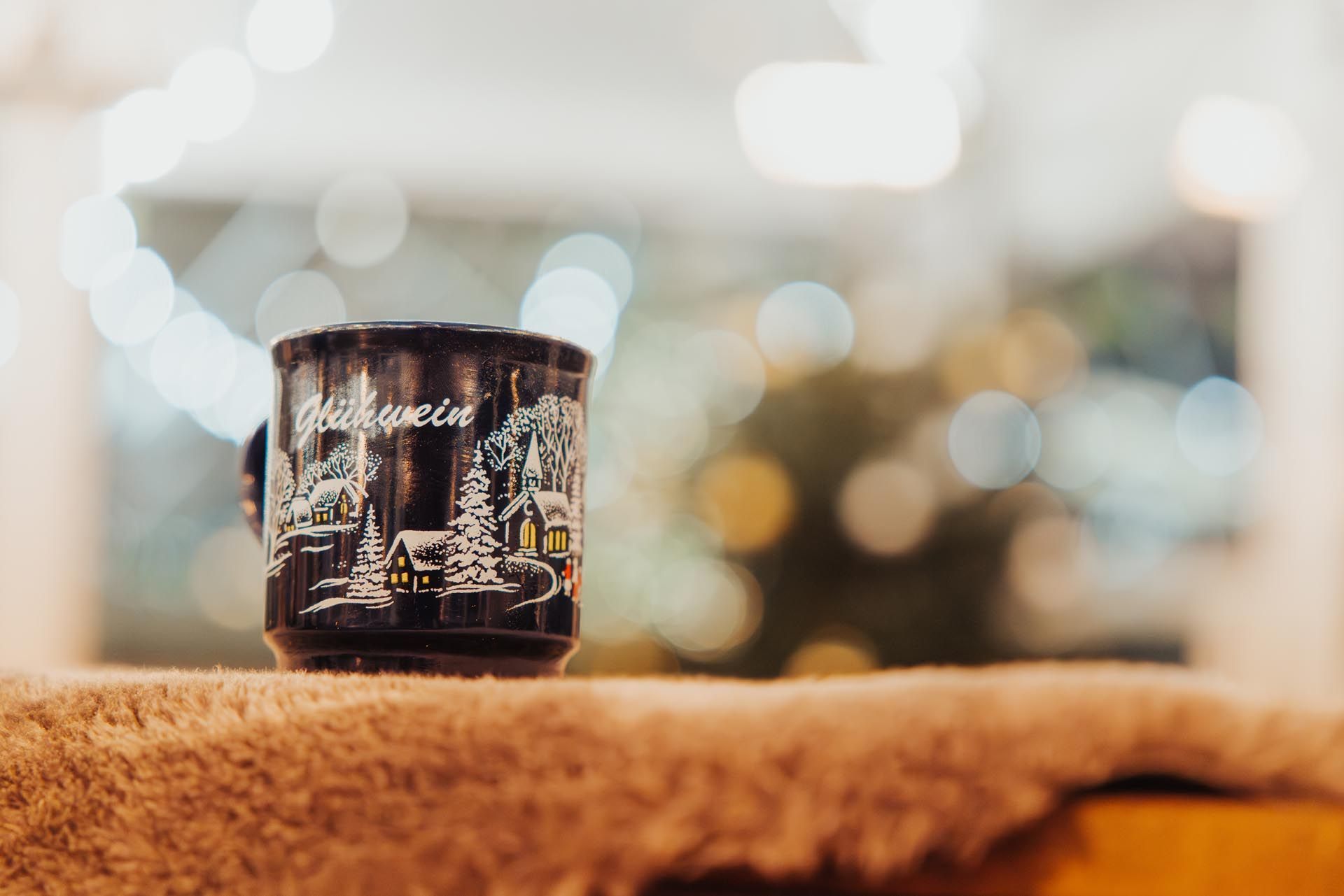 Dunkle Tasse mit Wintermotiv auf einer flauschigen Decke, im Hintergrund verschwommenes Licht.