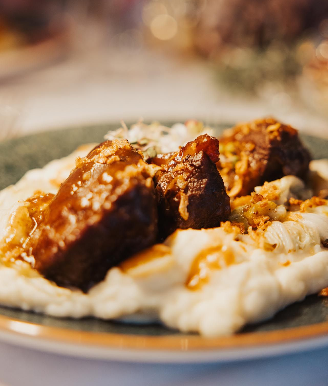 Geschmorte Rinderrippchen auf Blumenkohlpüree, mit Sauce beträufelt, auf einem Teller mit grünem Rand.