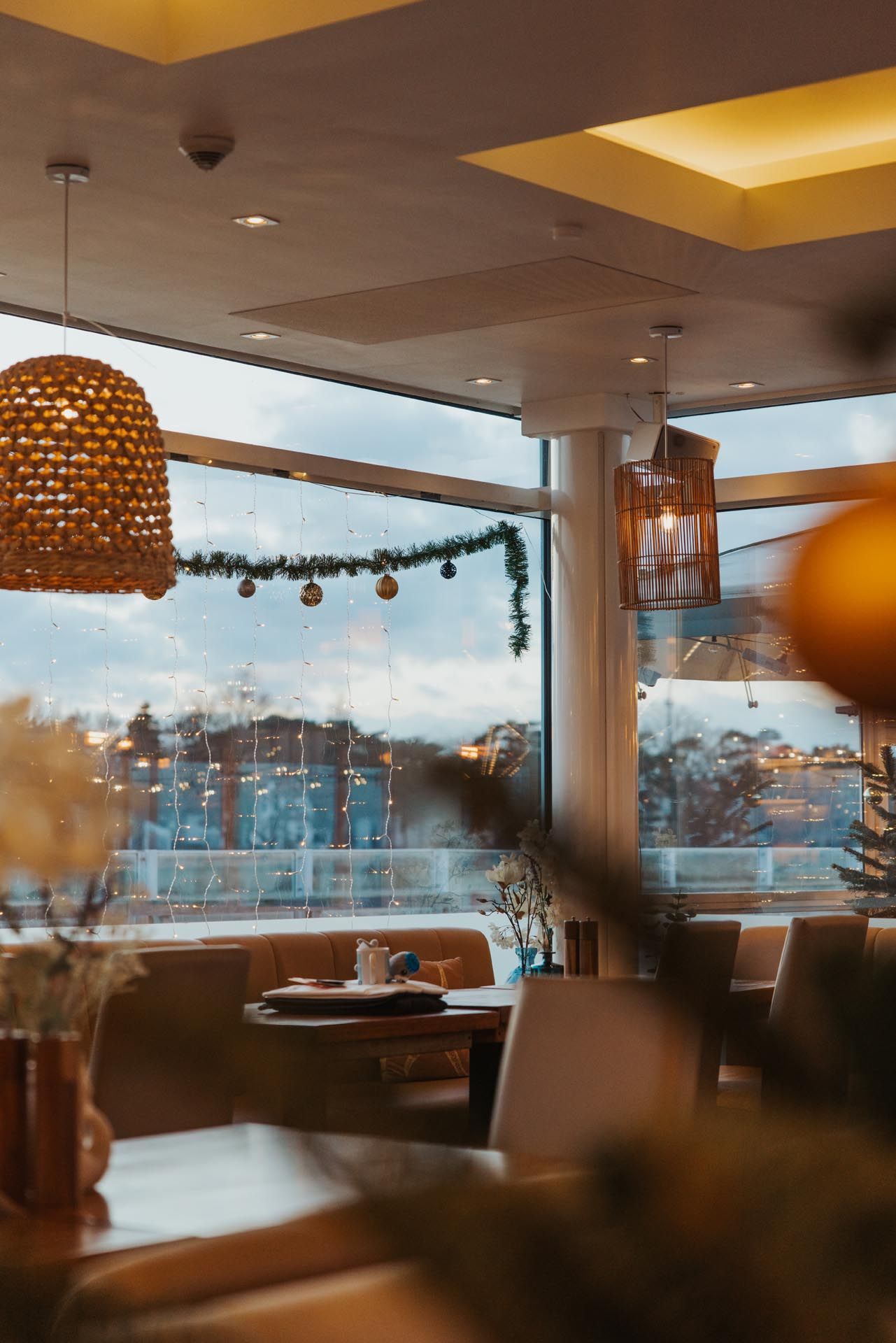 Innenansicht eines Restaurants mit Korblampe, einem Fenster mit Blick auf die Stadtlandschaft und Tischen.