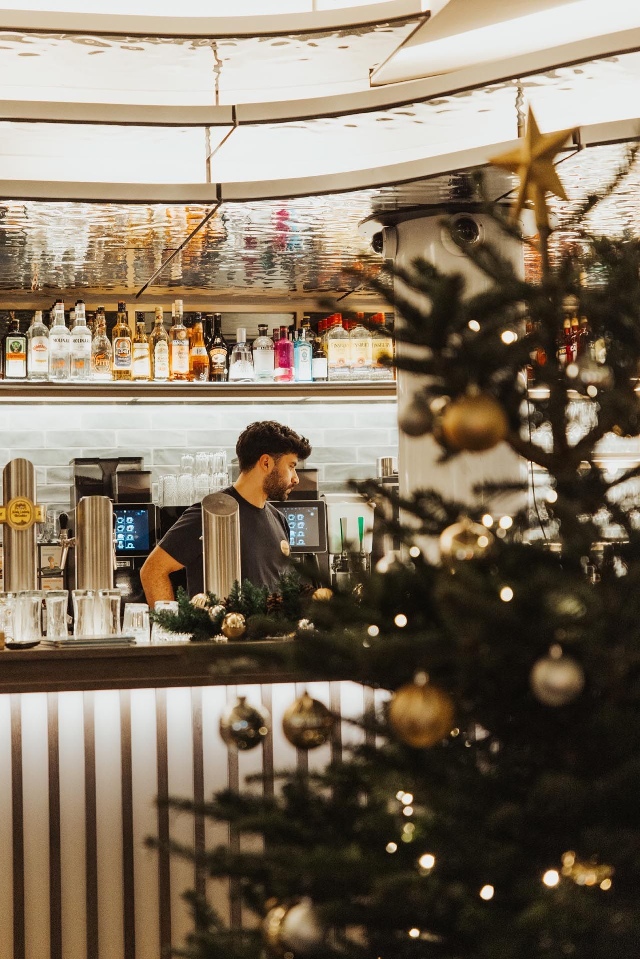 Barkeeper an der Bar mit einem Weihnachtsbaum davor. Flaschen mit Spirituosen auf der Rückwand der Bar.