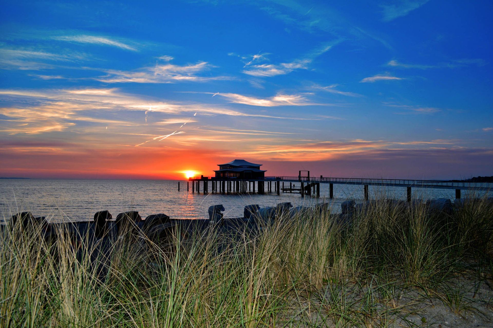 Sonnenuntergang über einem Pier mit einem Gebäude am Ende, Strandhafer im Vordergrund, leuchtende Blau- und Orangetöne.