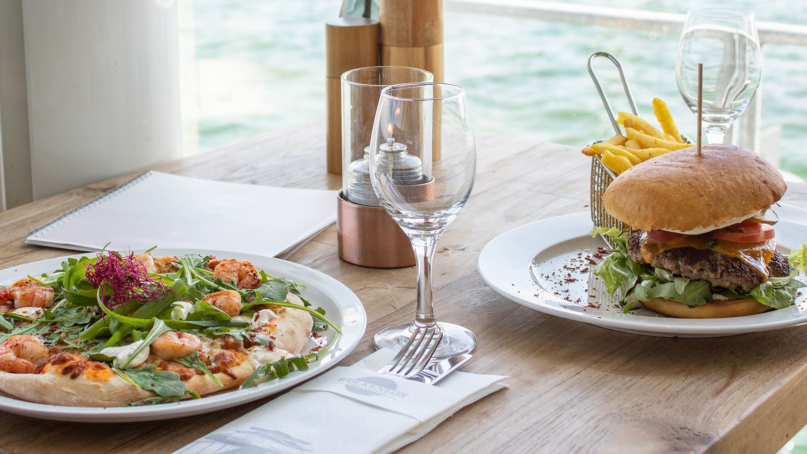 Pizza und Burger mit Pommes auf einem Holztisch in der Nähe von Wasser. Gläser und Servietten sind ebenfalls vorhanden.