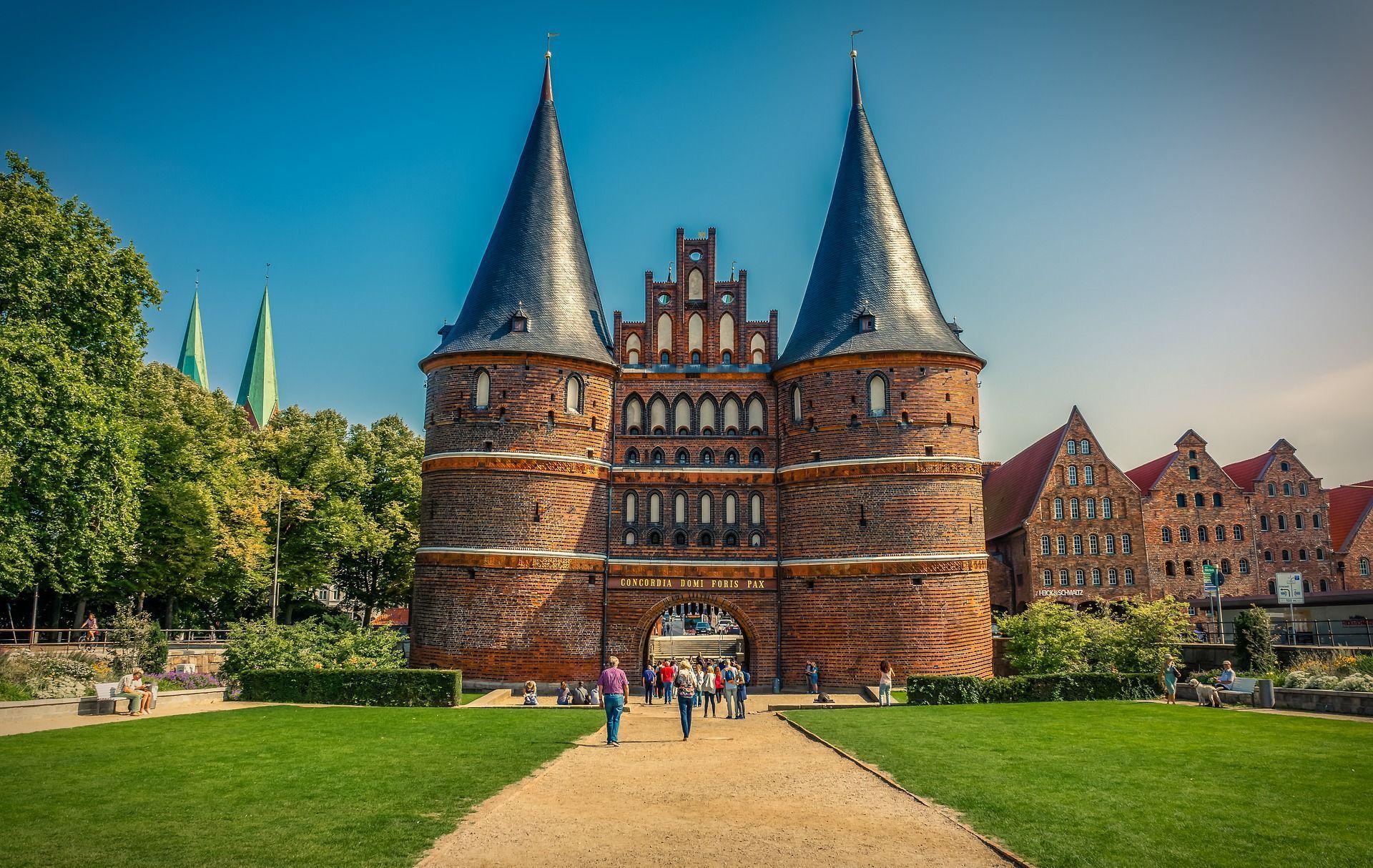 Burgtor, Lübeck, Deutschland, mit zwei hohen Türmen und einem gewölbten Eingang. Menschen gehen über eine grüne Wiese auf das Tor zu.