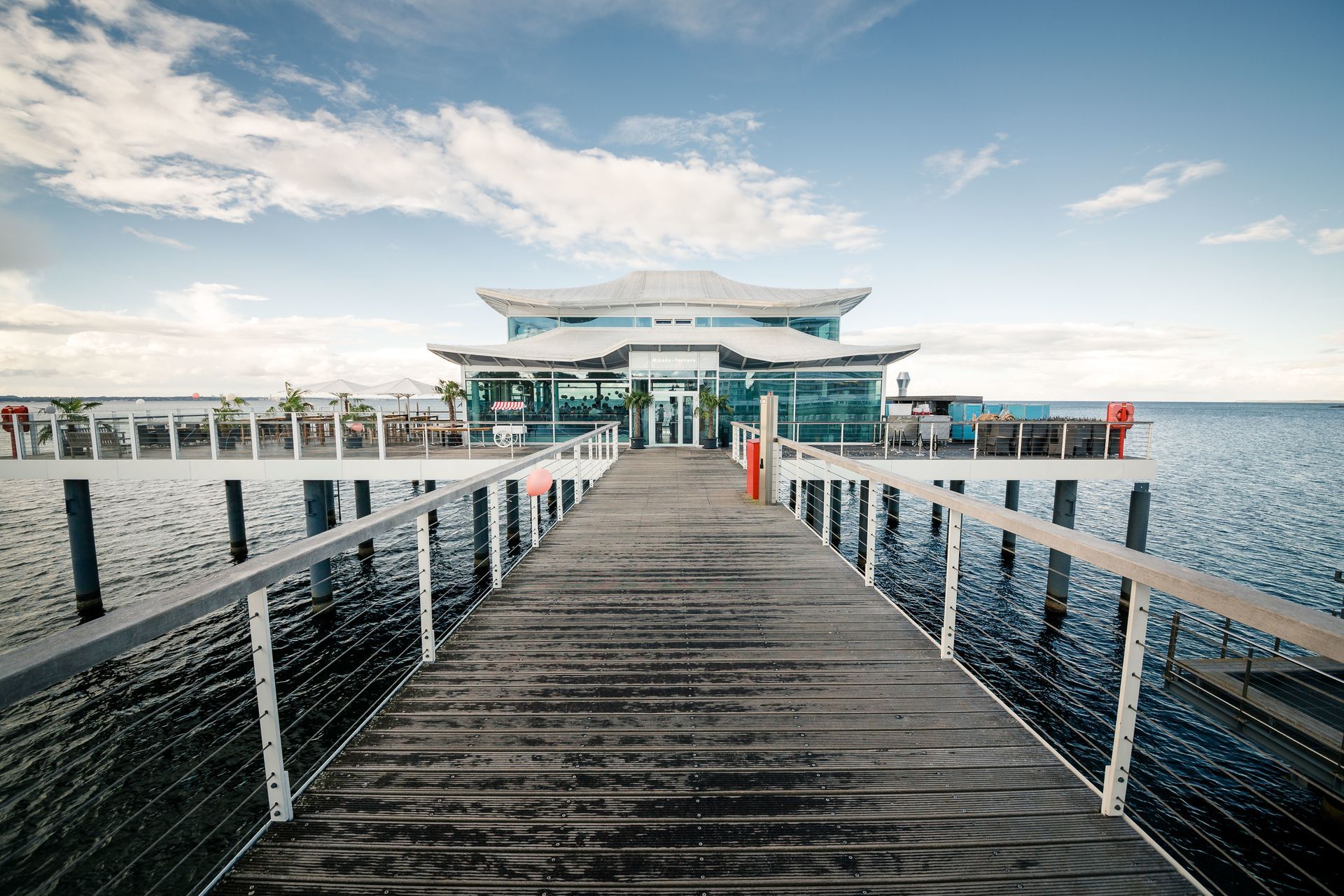 Holzsteg, der zu einem Gebäude über Wasser führt, blauer Himmel und Wolken darüber.