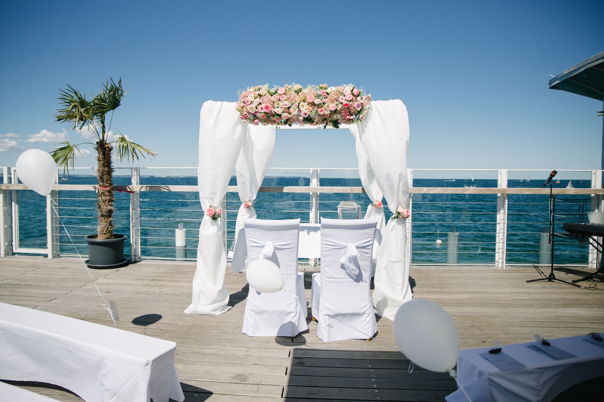 Hochzeitszeremonie auf einem Pier mit Blick auf den Ozean. Weiße Stühle, mit Blumen geschmückter Bogen, Luftballons.
