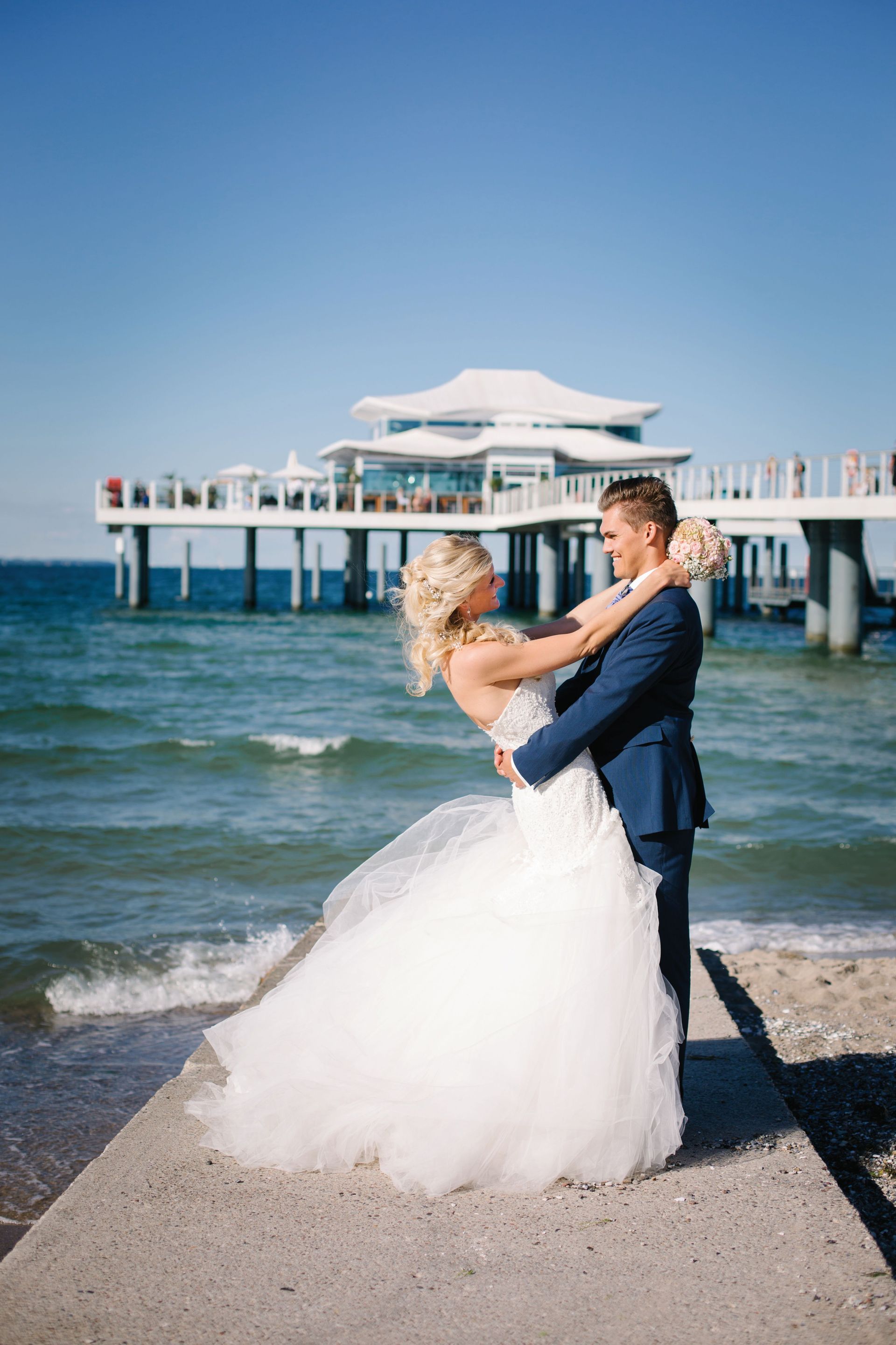 Frischvermählte umarmen sich auf einem Pier am Strand; Braut im weißen Kleid, Bräutigam im blauen Anzug, strahlend sonniger Tag.