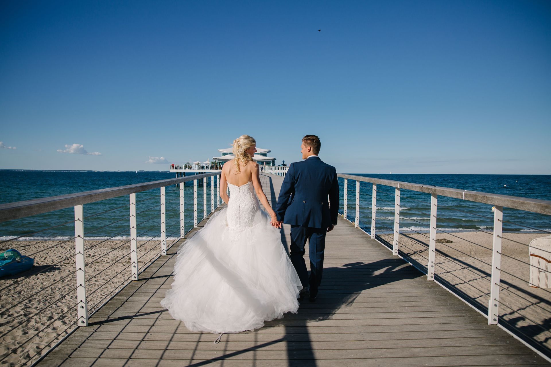 Braut und Bräutigam gehen an einem sonnigen Tag Hand in Hand auf einem Pier in Richtung Meer.