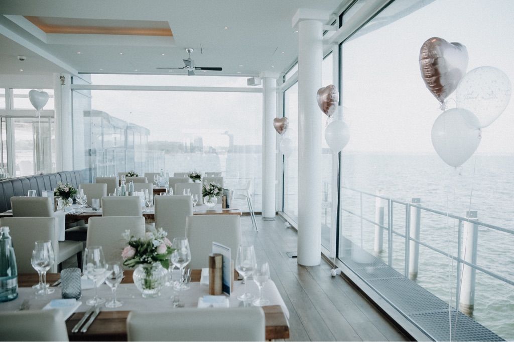 Eleganter Speisesaal mit für eine Veranstaltung gedeckten Tischen und Blick auf das ruhige Meer. Weiße und roségoldene Luftballons.