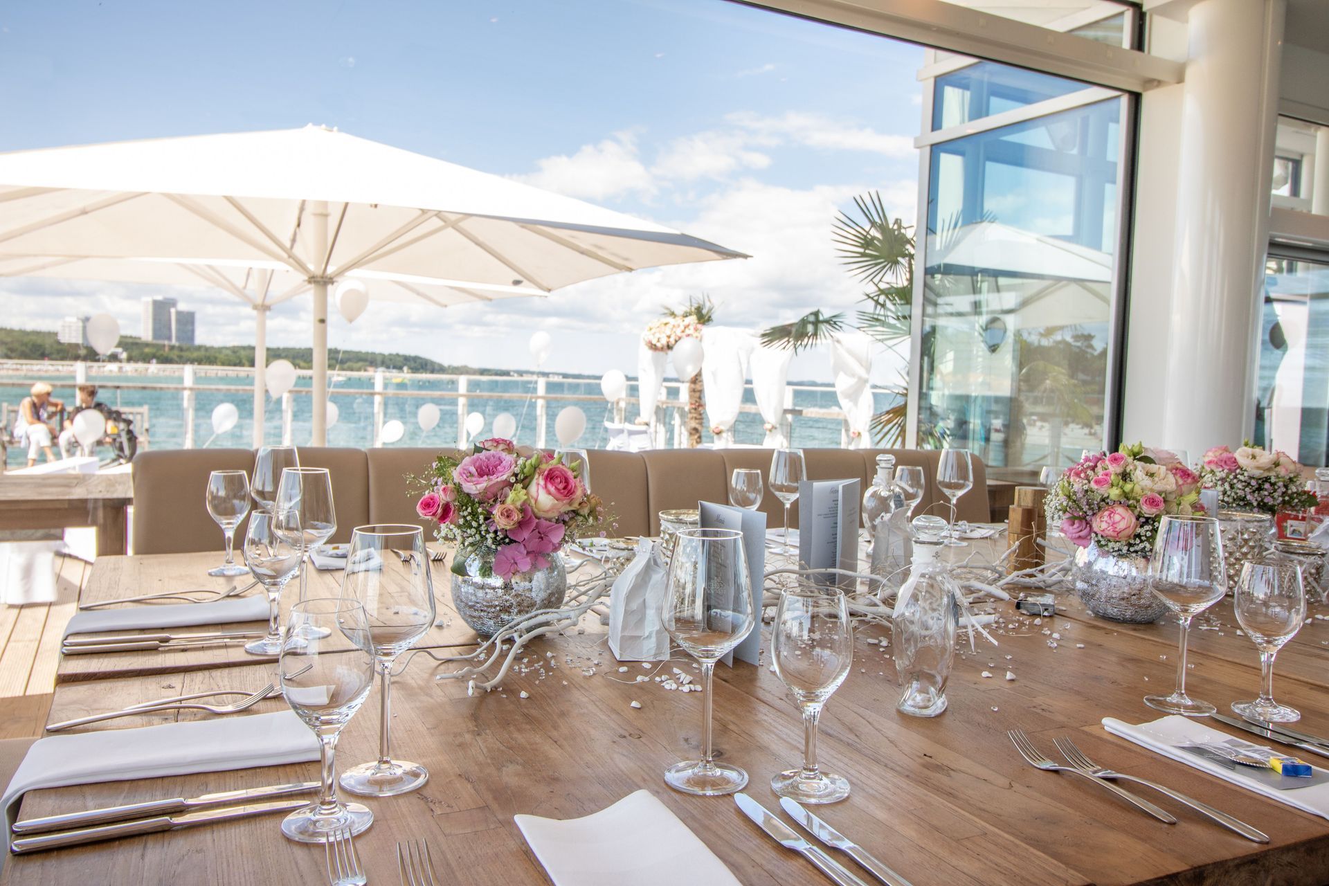 Gedeckter Tisch für eine Mahlzeit auf einer Terrasse am Wasser; Blumen, Glaswaren und Silberbesteck sind ausgestellt.