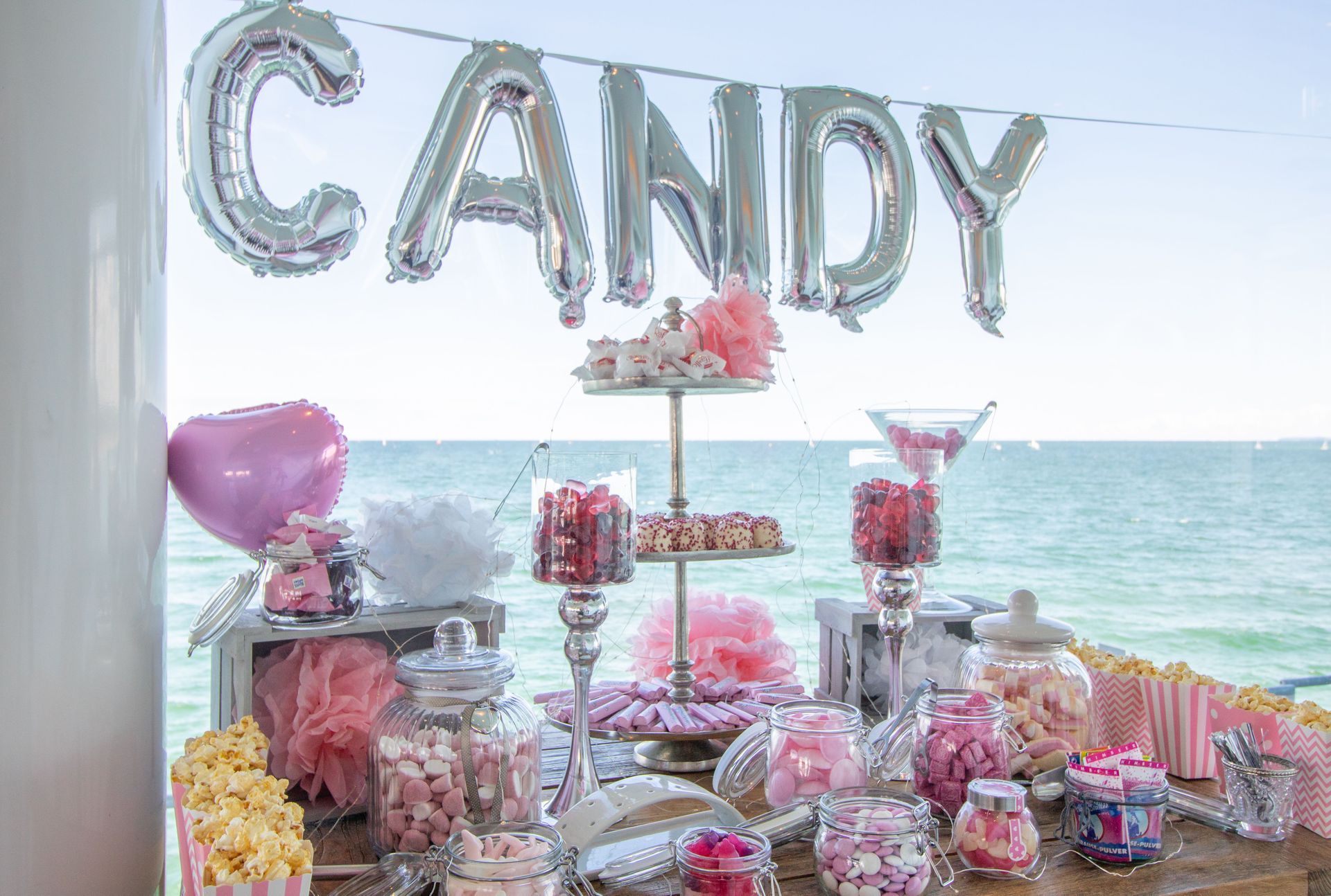 Süßigkeitenbuffet mit rosa und roten Leckereien, Luftballons und einer Meereskulisse.