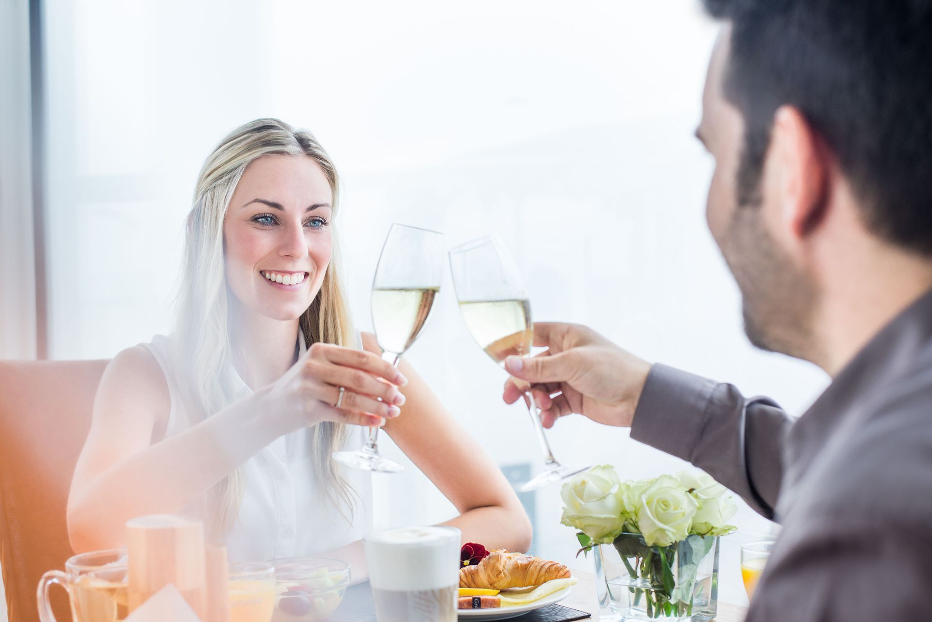 Ein Paar stößt an einem Tisch mit Champagnergläsern an. Die Frau lächelt, Sonnenlicht erhellt die Szene.