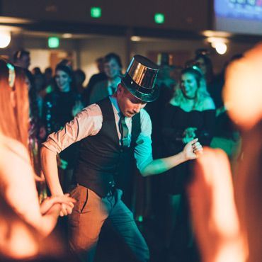 Mann mit Zylinder tanzt auf einer Party, andere schauen zu, bunte Beleuchtung.
