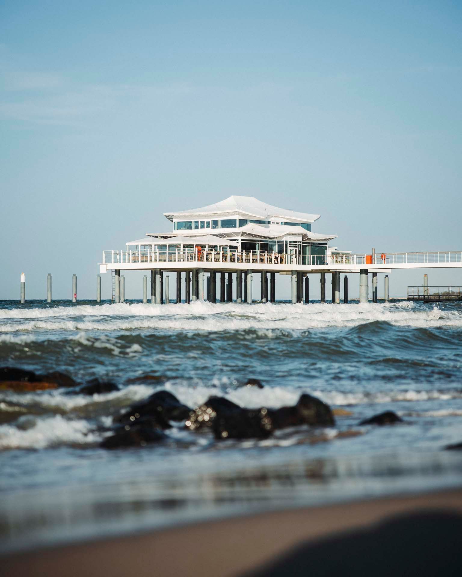 Pier, der in den Ozean hineinragt, weißes Gebäude mit einem Restaurant darauf. Wellen brechen an der Küste, blauer Himmel.