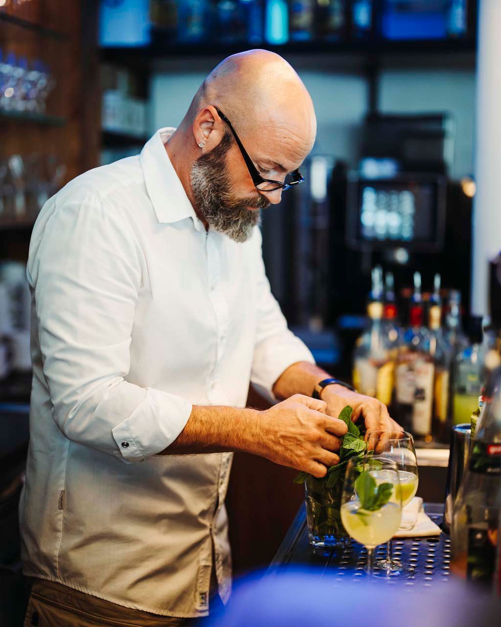 Barkeeper mit weißem Hemd und Bart gibt in einer Bar Minze in einen Cocktail.