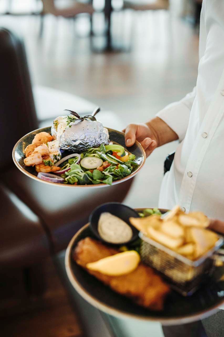 Person hält zwei Teller mit Essen: Fish and Chips, Salat mit Lachs und eine Ofenkartoffel.
