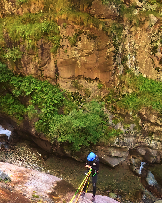 Persona haciendo rápel por un acantilado rocoso cubierto de vegetación, con un arroyo debajo.