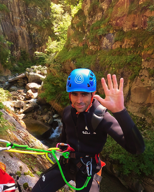 Hombre con traje de neopreno y casco descendiendo en rapel por un cañón, saludando. Paredes verdes del cañón y agua al fondo.