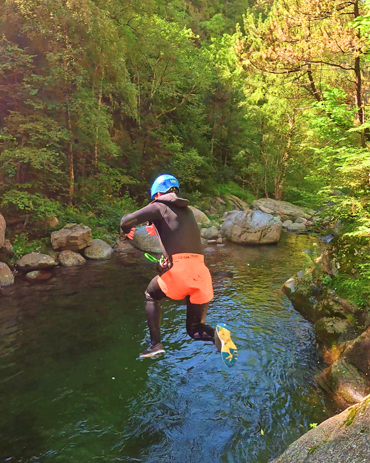 Persona saltando a un río verde, vistiendo traje de neopreno y casco, rodeado de árboles y rocas.