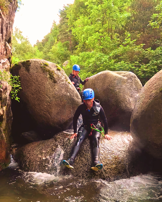 Dos personas con trajes de neopreno y cascos sobre rocas mojadas en un arroyo, rodeadas de árboles.