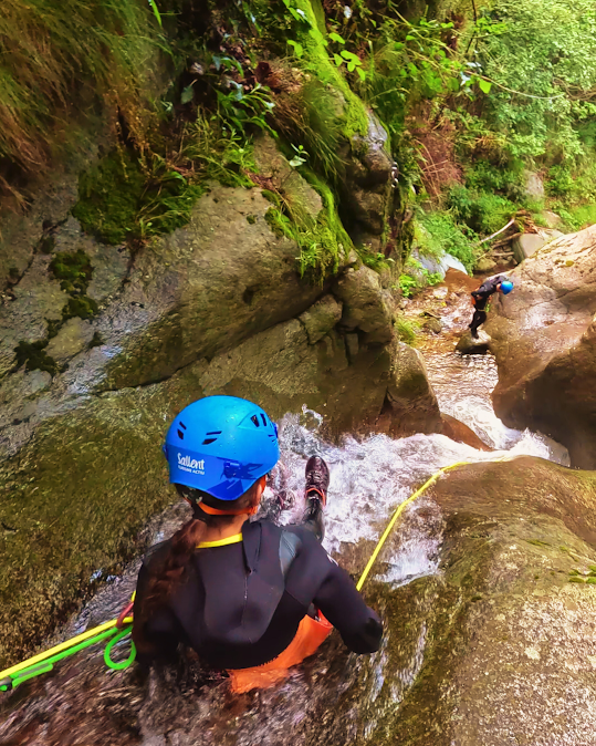 Persona haciendo rápel por una cascada con equipo de barranquismo, otra persona visible al fondo.