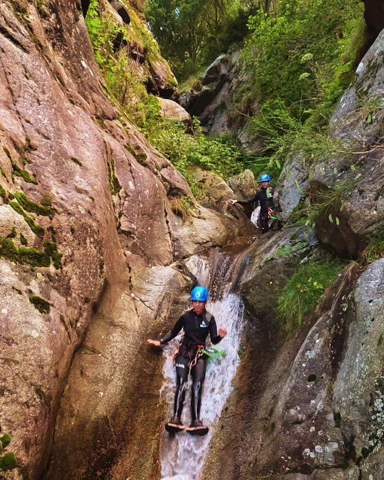 Dos personas hacen barranquismo por una estrecha cascada. Llevan casco, traje de neopreno y una tabla. Una exuberante vegetación rodea las rocas.