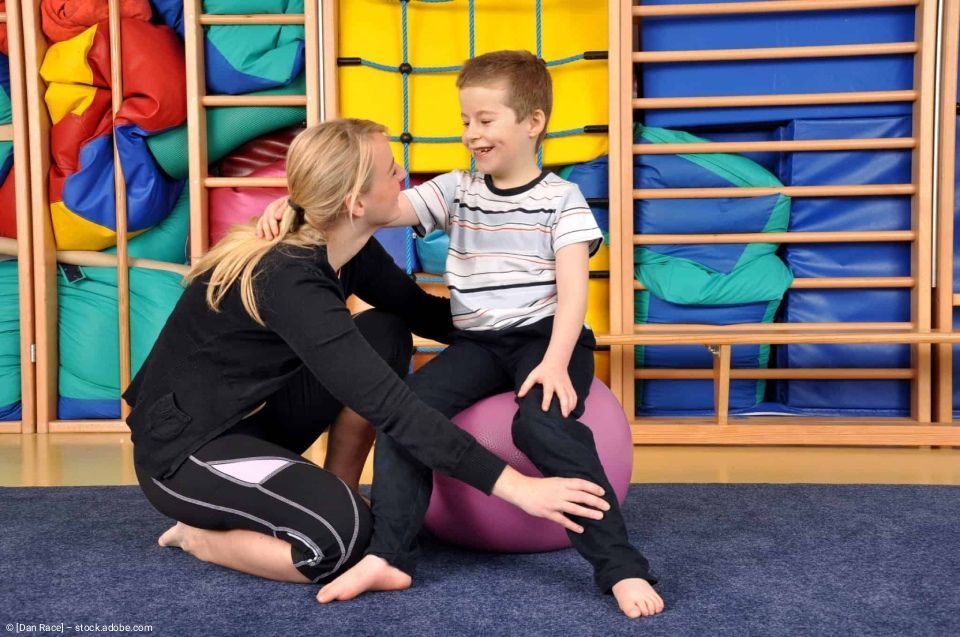 Eine professionelle Therapeutin unterstützt ein Kind, das in einer farbenfrohen Turnhalle auf einem lila Gymnastikball sitzt.