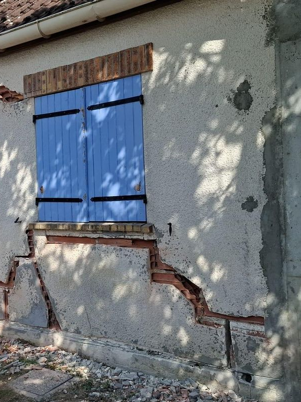 Stuc fissuré sur la façade d'une maison, laissant apparaître des briques apparentes. Des volets bleus et un linteau en bois surplombent la fenêtre.