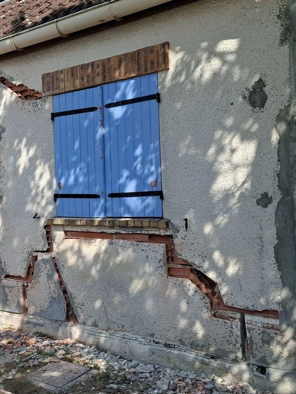 Extérieur d'un bâtiment avec des volets bleus et du stuc fissuré révélant la maçonnerie.