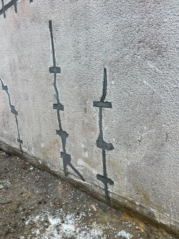 Mur en stuc gris avec fissures réparées et mastic foncé. La base rencontre le sol humide.