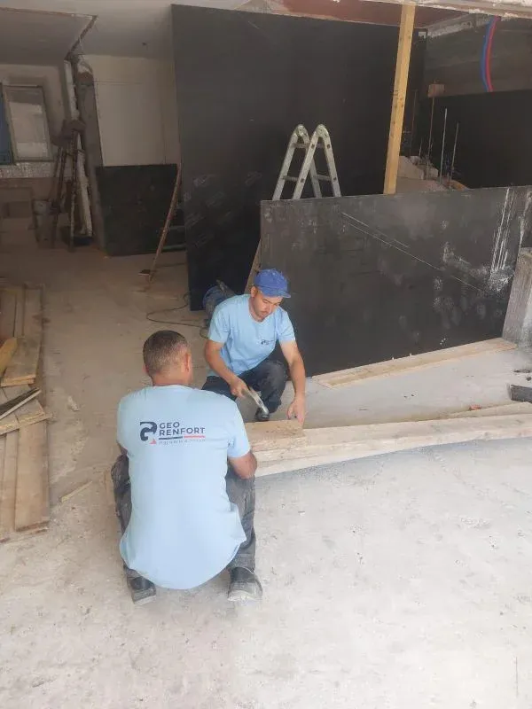 Deux hommes, vêtus de chemises bleues, travaillent le bois dans un atelier. Derrière eux, des panneaux noirs, une échelle à proximité.