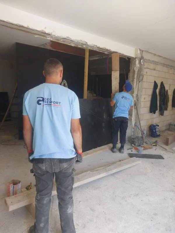 Deux hommes en chemise bleue travaillent sur un chantier. L'un se tient face à un mur noir, l'autre travaille dans une ouverture.