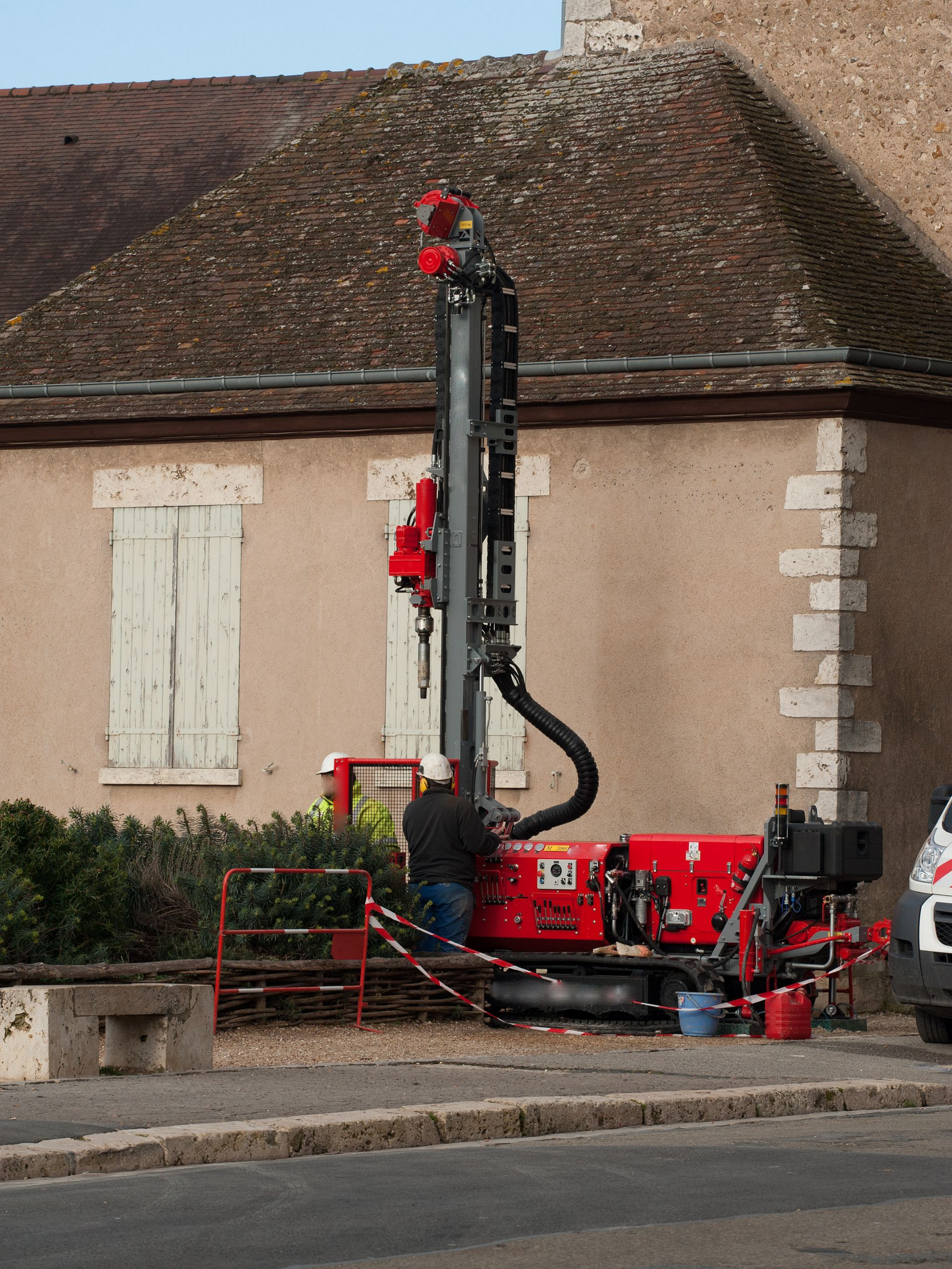 Une perceuse rouge près d'un bâtiment ; un ouvrier en vêtements sombres se tient à proximité, préparant l'équipement.