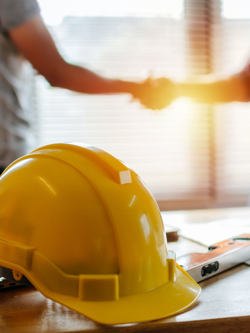 Casque jaune sur une table, deux personnes se serrant la main en arrière-plan, intérieur ensoleillé.